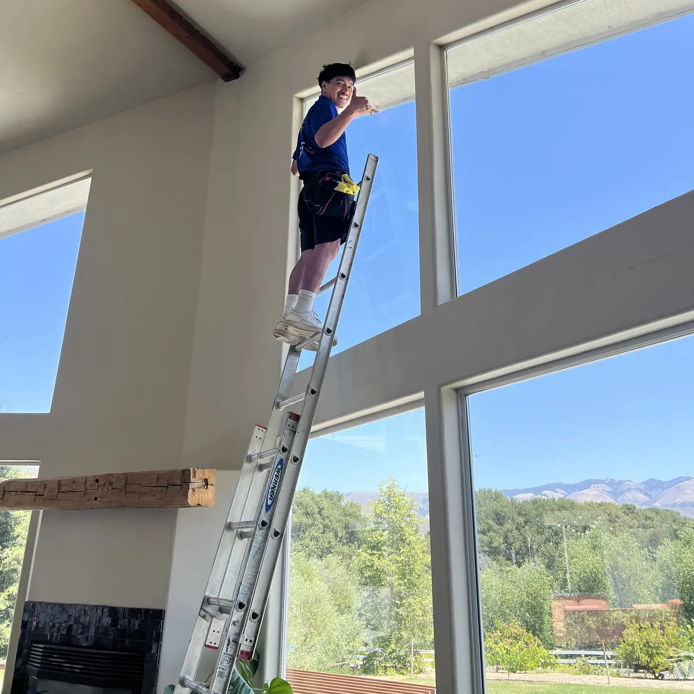 High Tide crew member on a ladder cleaning tall living-room windows with Central Coast hills outside